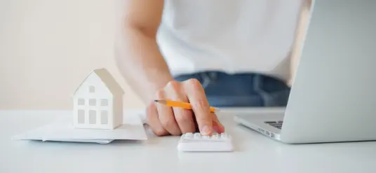 Mann benutzt am Laptop Haushaltsrechner von 4baufi, daneben ein Taschenrechner und ein Hausmodell